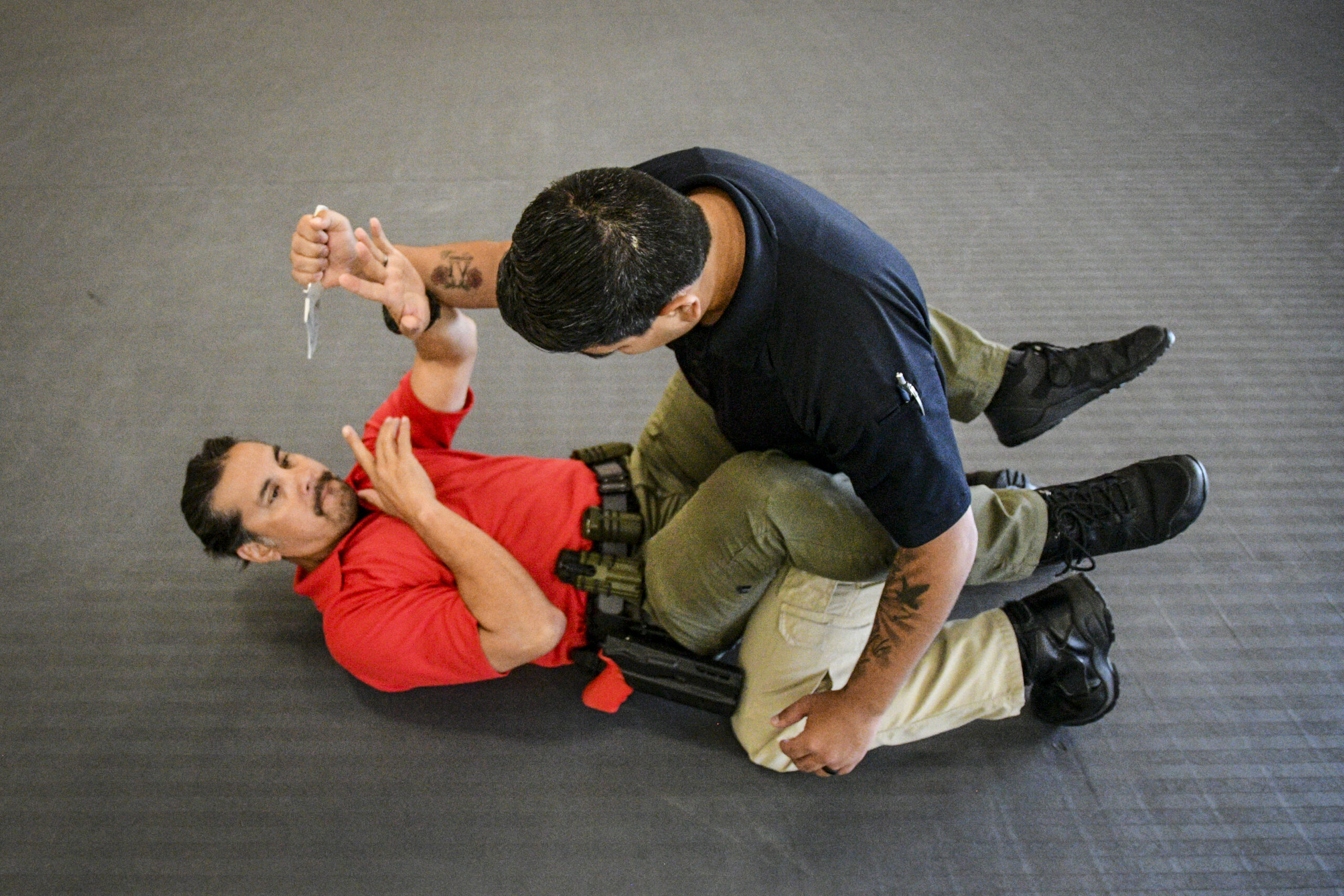 Two men fighting on the ground; the man on top is holding a knife and the man on the bottom is engaging in ground SERE techniques
