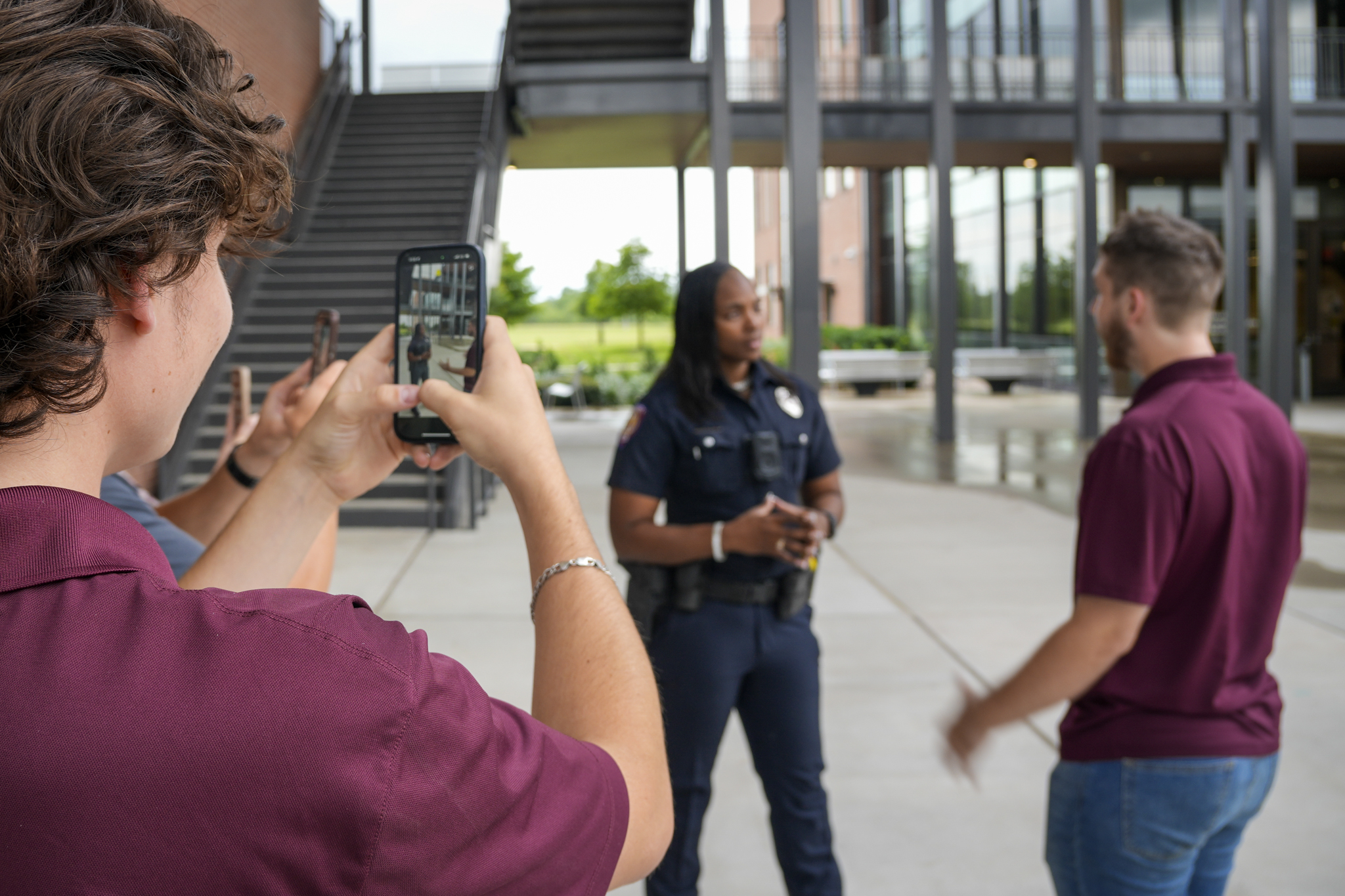 A young male is speaking with a law enforcement officer in a public setting. Two bystanders are recording the interaction with their cell phone.