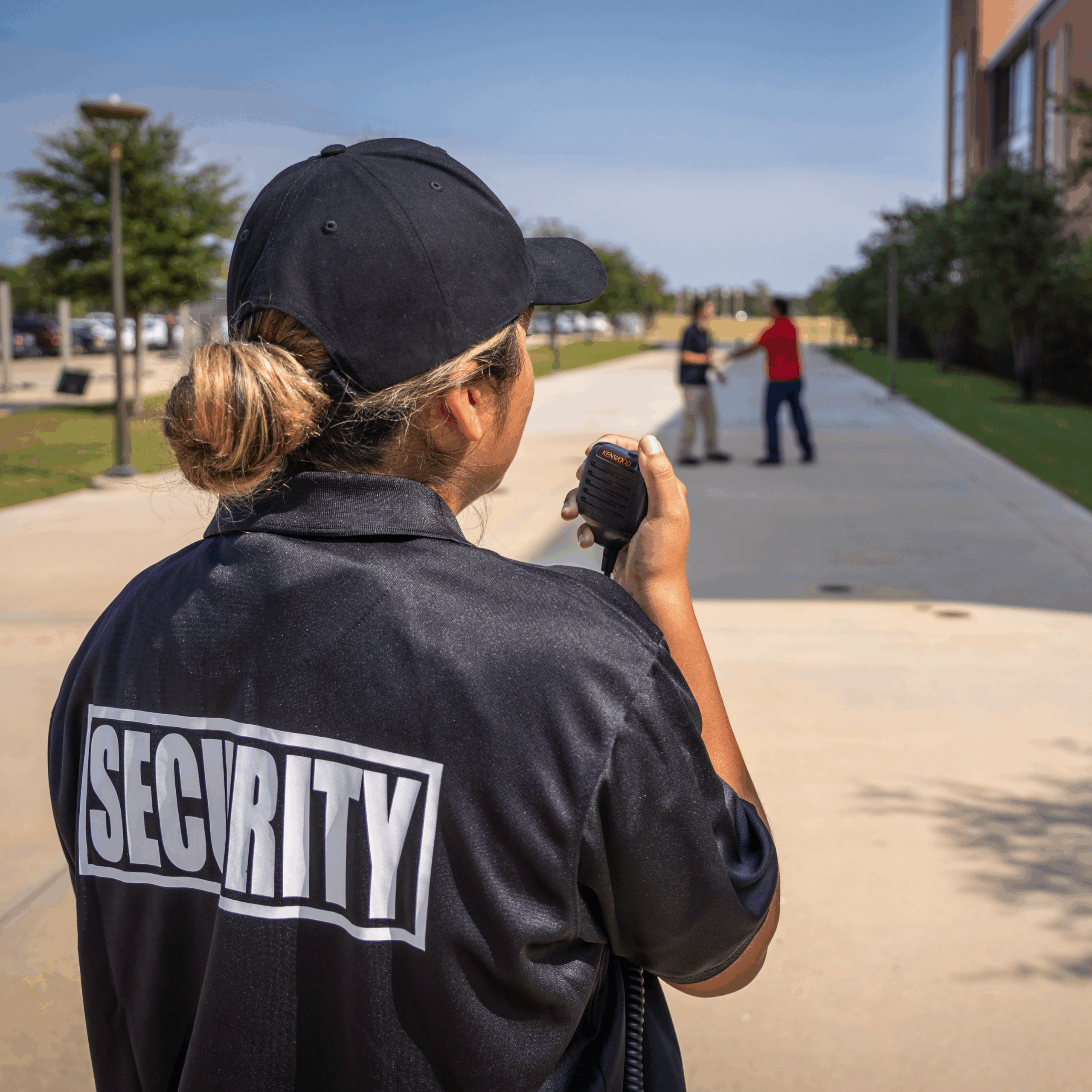 Female security officer calling in an incident over the radio outside of an office building with two individuals involved in a possible altercation in the distance