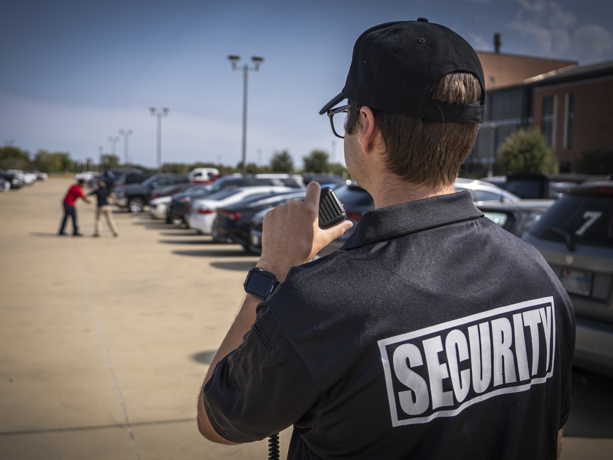 A security guard in a parking lot calling for backup on a two-way radio while two individuals are fighting in the distance.